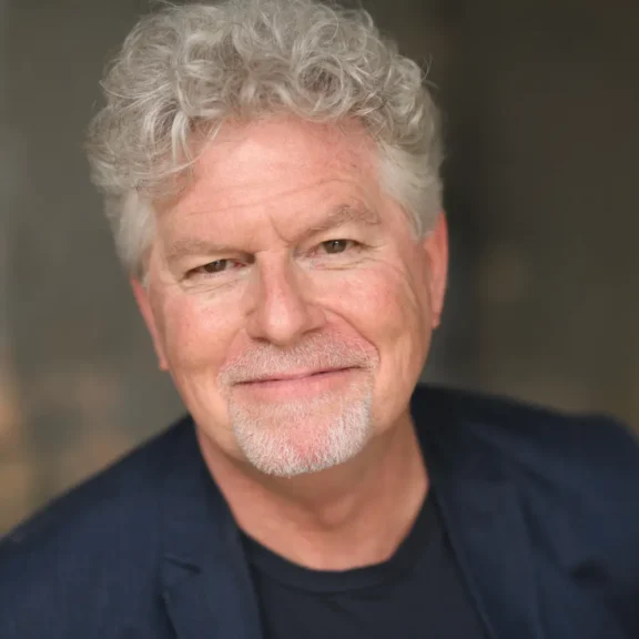 A middle-aged man with curly gray hair and a goatee, wearing a dark jacket and black shirt, smiles slightly at the camera against a blurred neutral background.