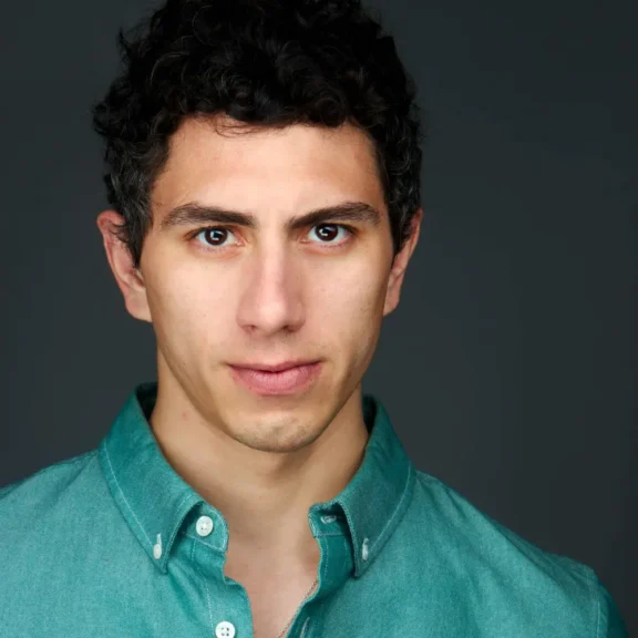 A young man with short curly dark hair and light skin wears a teal button-up shirt and looks directly at the camera against a plain dark background.