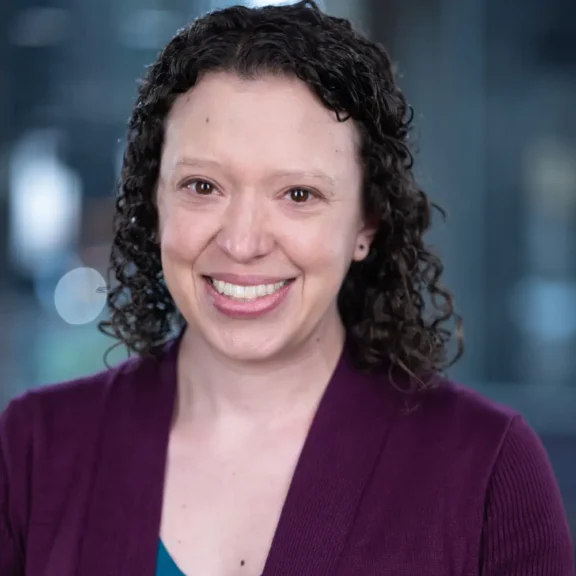 A woman with curly brown hair, wearing a purple cardigan and teal top, smiles at the camera against a blurred indoor background.