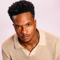 A man with short curly hair and a trimmed beard wears a beige knit collared shirt and gold chain, facing the camera against a light background.