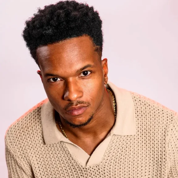 A man with short curly hair and a trimmed beard wears a beige knit collared shirt and gold chain, facing the camera against a light background.
