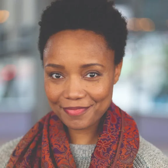 A woman with short curly hair, wearing a patterned red scarf and gray sweater, looks at the camera and smiles softly. The background is softly blurred.