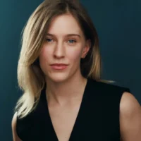 A woman with straight, shoulder-length blonde hair and blue eyes is wearing a sleeveless black top and looking directly at the camera against a dark background.
