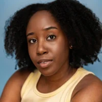 A woman with natural curly hair, wearing a sleeveless yellow top, looks directly at the camera against a light blue background.