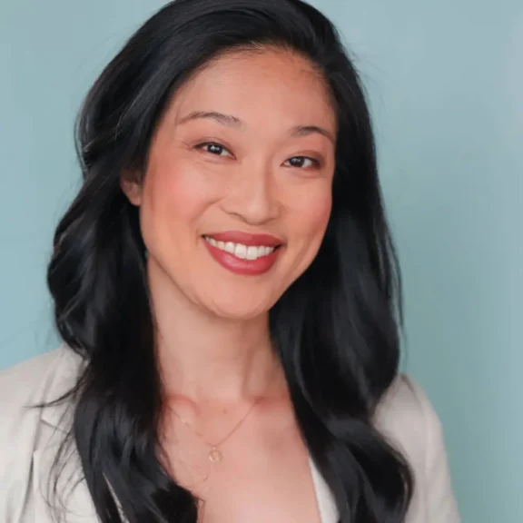 Woman with long black hair and a light blazer smiles at the camera against a plain light blue background.