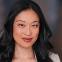A woman with long black hair and light makeup smiles softly at the camera against a blurred, neutral background.
