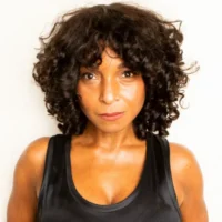 A woman with short, curly dark hair wearing a black sleeveless top stands against a plain light background, looking directly at the camera.