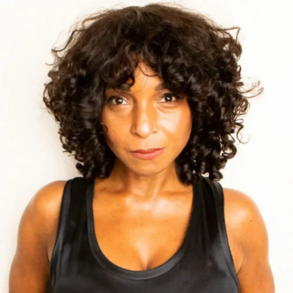 A woman with short, curly dark hair wearing a black sleeveless top stands against a plain light background, looking directly at the camera.