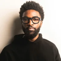 A man with a beard and afro hairstyle, wearing black glasses and a black hoodie, stands against a plain light-colored background.