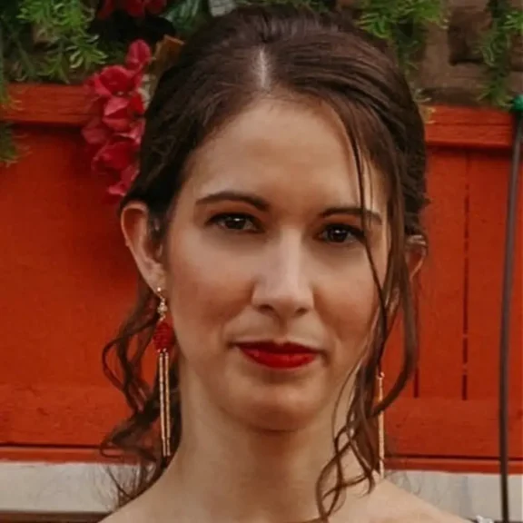 A woman with dark brown hair, red lipstick, and gold earrings stands in front of an orange wooden fence decorated with greenery and red flowers.