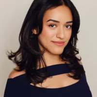 A woman with dark shoulder-length hair, wearing a navy blue off-the-shoulder top, stands against a plain light background and looks at the camera with a slight smile.
