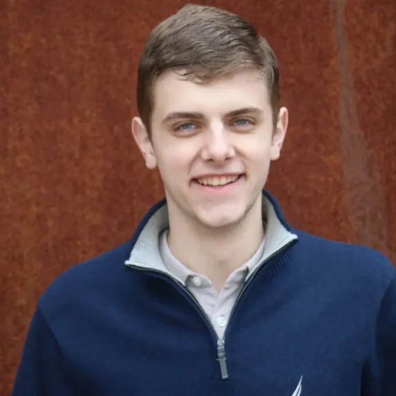 A young man with short brown hair and blue eyes is smiling at the camera, wearing a navy zip-up sweater over a light gray collared shirt, standing in front of a rust-colored background.