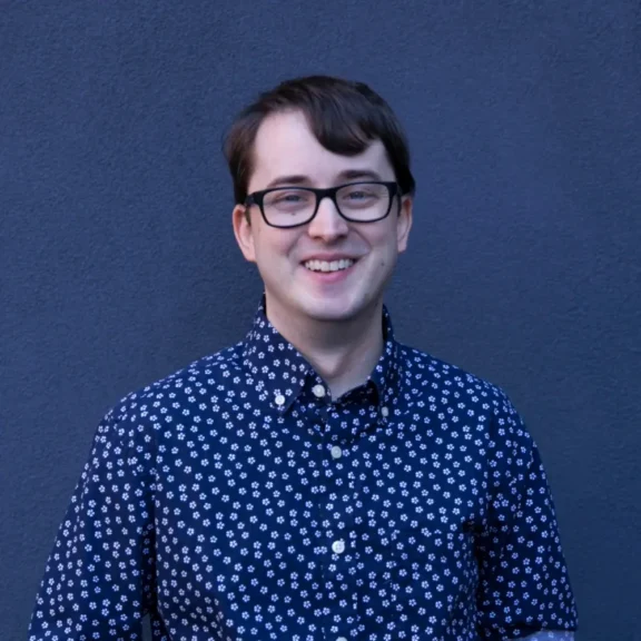 A person with short brown hair and glasses, wearing a blue shirt with a white floral pattern, stands in front of a plain dark blue wall, smiling at the camera.