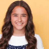 A girl with long wavy brown hair smiles at the camera. She is wearing a white top with a dark blue patterned dress, standing in front of a plain yellow background.