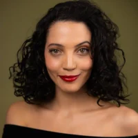 A woman with curly dark hair, red lipstick, and hoop earrings poses against a plain olive background, wearing an off-the-shoulder black top.