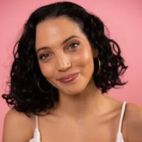Woman with short curly black hair, wearing hoop earrings and a white top, smiling softly against a pink background.