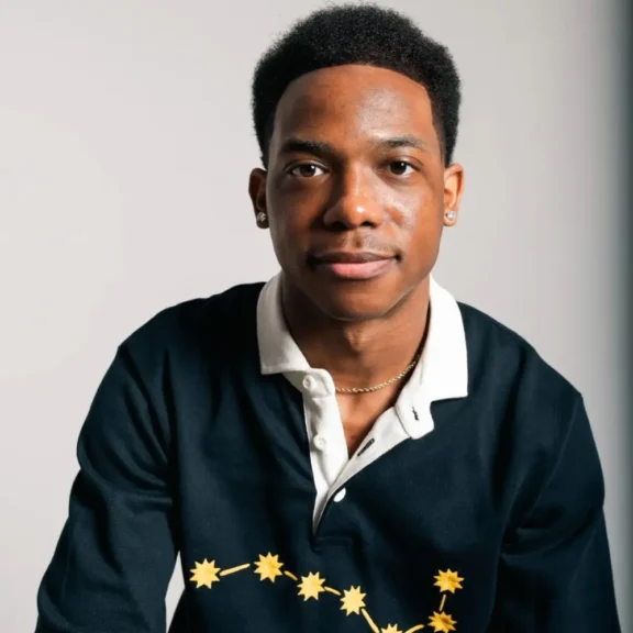A young man with short hair and earrings sits facing the camera, wearing a collared shirt with yellow star graphics on a dark background.