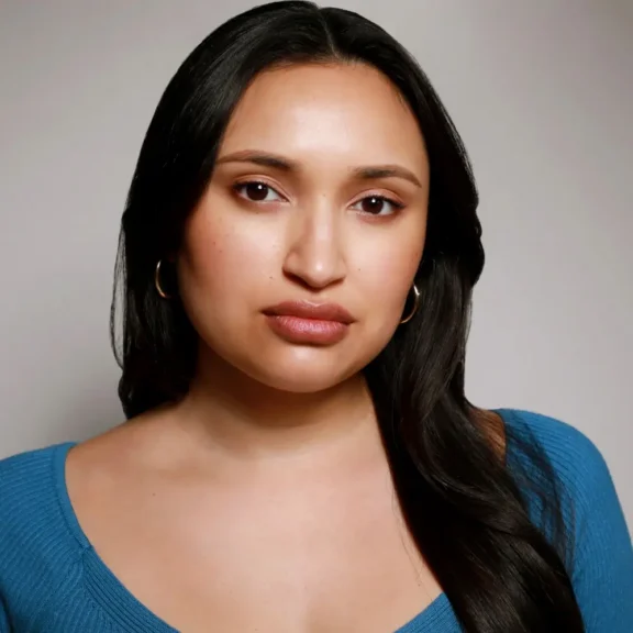 Woman with long dark hair wearing gold hoop earrings and a blue top, facing the camera against a neutral background.