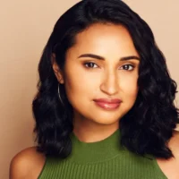 Woman with medium-length curly black hair, wearing a sleeveless green top and hoop earrings, posing against a beige background.