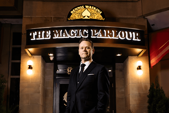 Dennis Watkins dressed in suit and tie stands in front of lit up "The Magic Parlour" sign.