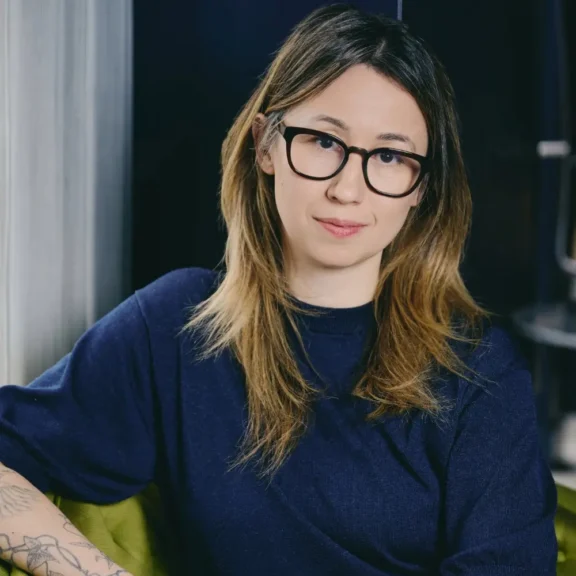 A woman with long light brown hair, glasses, and a dark blue top sits indoors, looking at the camera with a neutral expression.