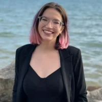 A person with shoulder-length brown and pink hair, wearing glasses, a black top, and a black blazer, smiles while standing outdoors near a body of water.