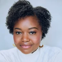 A woman with short, curly hair is wearing a white sweater and hoop earrings, looking directly at the camera against a light background.