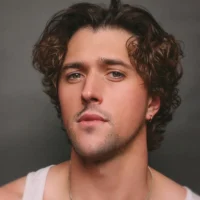 A man with wavy brown hair, light stubble, and an earring looks at the camera. He is wearing a white tank top and a necklace. The background is plain and dark.