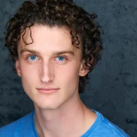 A young person with curly brown hair and blue eyes, wearing a light blue shirt, poses in front of a dark textured background.