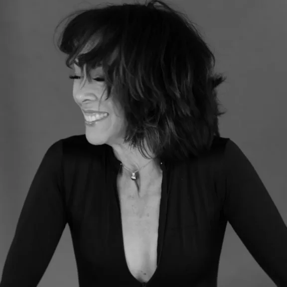 A woman with shoulder-length dark hair wearing a deep V-neck black top smiles and looks to her left against a plain backdrop.