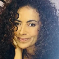 A woman with curly hair smiles softly while resting her chin on her hand, looking directly at the camera.