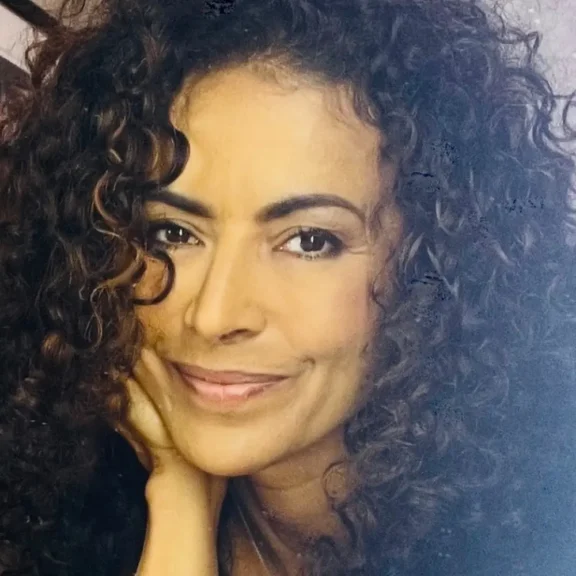 A woman with curly hair smiles softly while resting her chin on her hand, looking directly at the camera.
