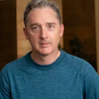 A man with short gray hair wearing a blue sweater looks directly at the camera against a brown and gold blurred background.