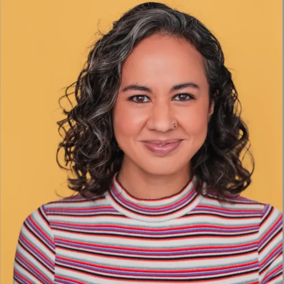 Woman with curly dark hair and a nose ring smiles at the camera, wearing a striped turtleneck, standing against a solid yellow background.