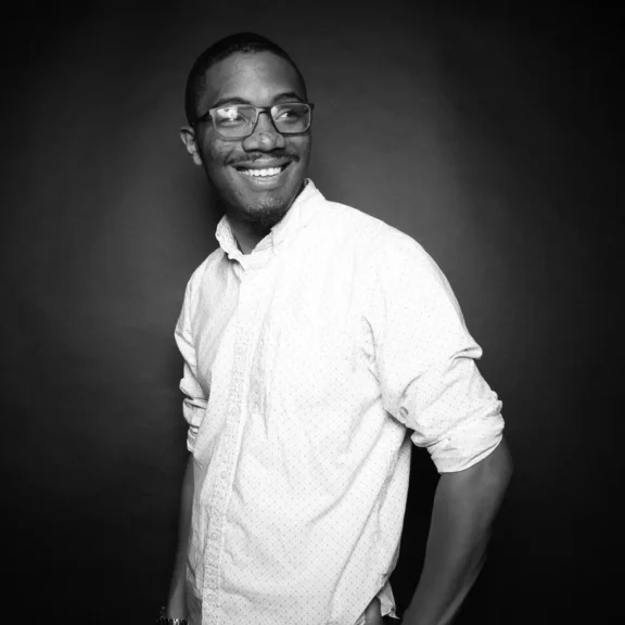 A man wearing glasses and a light-colored, long-sleeve shirt stands against a dark background, smiling and looking slightly to the side.