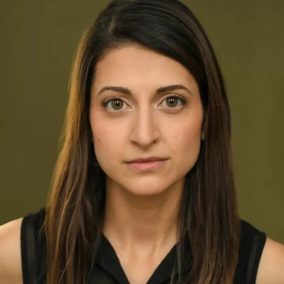 A woman with long brown hair and brown eyes, wearing a sleeveless black top, looks straight at the camera with a neutral expression in front of an olive green background.