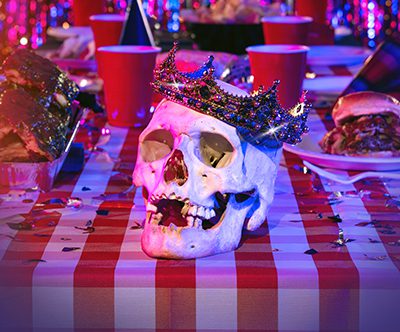 A jewel-encrusted crown adorns a skull sitting on a table littered with red Solo cups and barbecued meats.