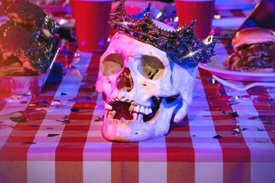 A jewel-encrusted crown adorns a skull sitting on a table littered with red Solo cups and barbecued meats.