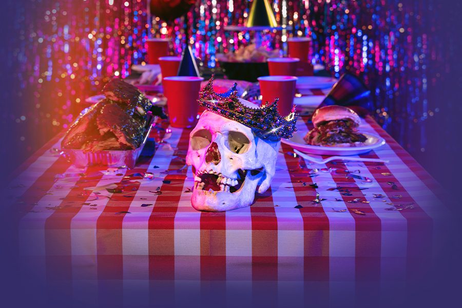 A jewel-encrusted crown adorns a skull sitting on a table littered with red Solo cups and barbecued meats.