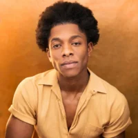 A person with short curly hair wearing a light yellow shirt sits against a warm, brown-toned background, looking directly at the camera.