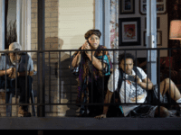 Three people sitting on a porch looking surprised while one is on the phone and another is recording an incident on the street.