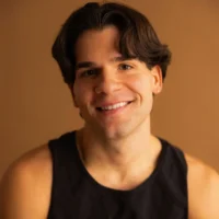 A young man with medium-length brown hair smiles at the camera, wearing a black sleeveless shirt against a plain brown background.