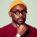 Person wears a yellow beanie, black rimmed glasses and maroon, rests head on fist.