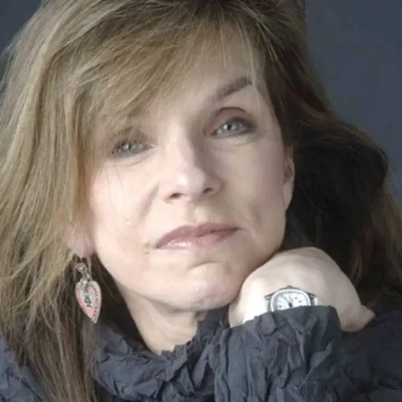 A woman with light brown hair rests her chin on her hand, wearing a watch and a heart-shaped earring, against a dark background.
