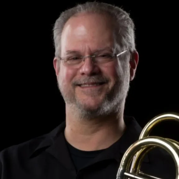 A middle-aged man with glasses and a beard smiles while holding a brass instrument against a black background.