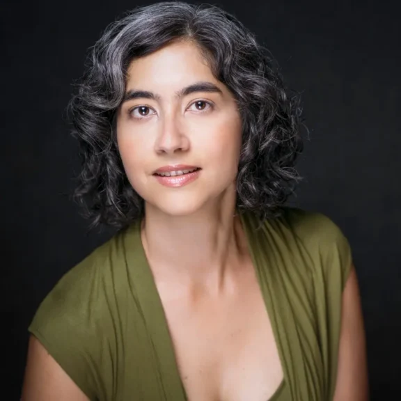 A woman with wavy salt-and-pepper hair, wearing an olive green top, poses in front of a dark background, looking directly at the camera.