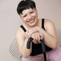 A person with short dark hair sits on a metal chair, smiling, and resting their arms on a black cane.