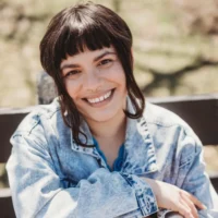 A person with medium-length dark hair and bangs, wearing a light denim jacket, sits outdoors on a bench, smiling at the camera.