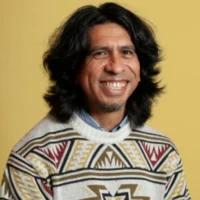 A man with medium-length dark hair, wearing a patterned sweater, smiles in front of a plain yellow background.