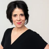 Woman with short dark hair, wearing a black V-neck top, looks at the camera with a slight smile against a plain light background.